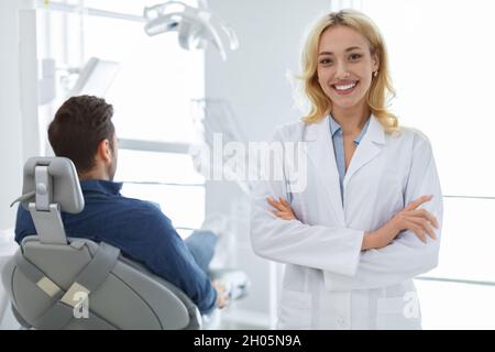 Jeune femme blonde souriante dentiste posant au beau blanc moderne cabinet dentaire, homme malade méconnaissable assis dans une chaise dentaire, en attente de salut Banque D'Images