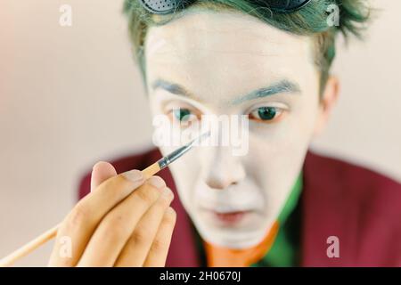MIM prépare le maquillage pour les performances.Concept de joueur de clown.Homme aux cheveux verts en costume bordeaux.Ambiance théâtre.L'acteur masculin couvre son visage par de la peinture à la greasepaint blanche à l'aide d'un pinceau. Banque D'Images