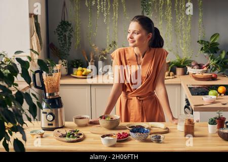 Bonne jeune femme dans une robe décontractée debout près d'une table de cuisine avec divers ingrédients pour smoothie maison Banque D'Images