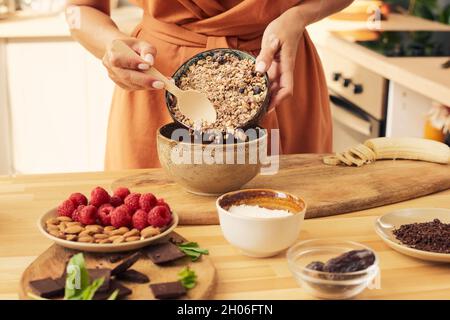 Mains de la femelle avec une cuillère en bois mettant le muesli dans le bol tout en préparant le dessert Banque D'Images