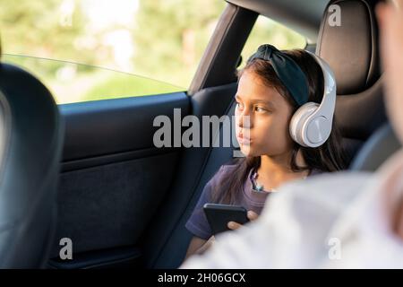 Jolie fille sérieuse avec casque défilant dans le smartphone tout en regardant à travers la fenêtre de la voiture Banque D'Images