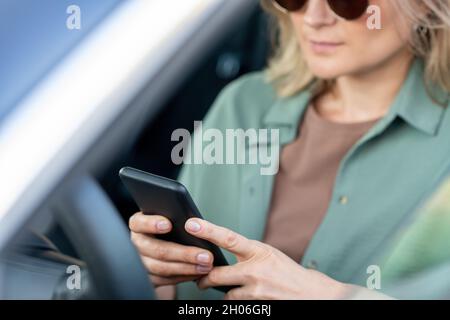 Mains de femme élégante défilant dans un téléphone intelligent tout en étant assis au volant dans la voiture Banque D'Images