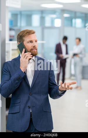 Homme d'affaires barbu en costume bleu communiquant sur smartphone contre deux collègues Banque D'Images