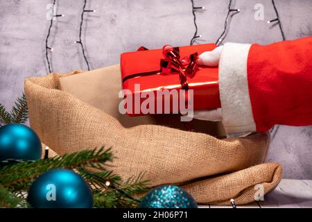 Le Père Noël prépare le sac de jute avec des cadeaux de Noël.Main avec gant blanc tenant une boîte cadeau avec papier et ruban rouge. Banque D'Images