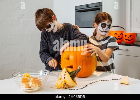 Deux jeunes enfants avec le visage de crâne de couper une citrouille dans leur cuisine. Banque D'Images