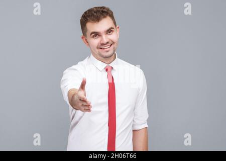 Portrait d'un jeune beau homme heureux en chemise blanche et cravate debout et regardant l'appareil photo avec un sourire crasseux et donnant la main à l'accueil ou à la poignée de main. Intérieur isolé sur fond gris. Banque D'Images