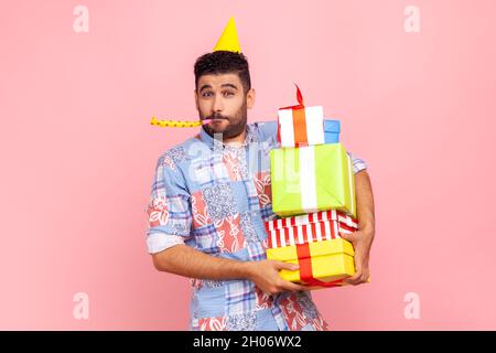 Portrait d'un homme joyeux et drôle avec une barbe portant une chemise et un cône de style décontracté, tenant une pile de cadeaux et un pavillon de fête soufflant, célébrant l'anniversaire.Studio d'intérieur isolé sur fond rose. Banque D'Images