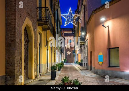 Vue sur la rue pavée étroite du soir parmi les anciens bâtiments avec éclairage de Noël à Alba, Piémont, Italie du Nord. Banque D'Images