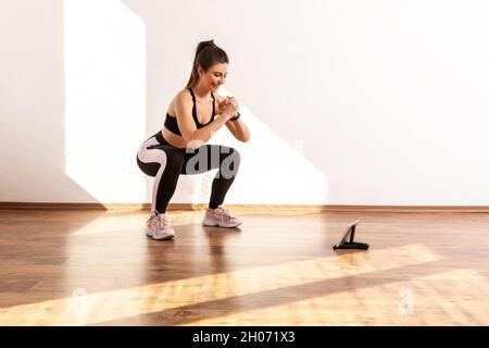 Femme sportive faisant des squats tout en regardant la classe de fitness en ligne, apprenant la technique correcte d'exercice, portant le haut de sport noir et des collants.Prise de vue en studio pleine longueur éclairée par la lumière du soleil depuis la fenêtre. Banque D'Images