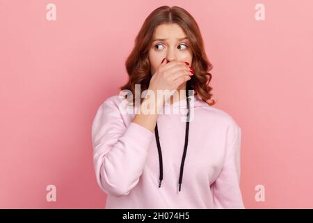 Portrait de la jeune fille de l'adolescence à poil dur choqué dans le sweat à capuche décontracté fermer la bouche avec la main, en gardant l'information secrète, Oh mon geste de dieu.Studio d'intérieur isolé sur fond rose Banque D'Images