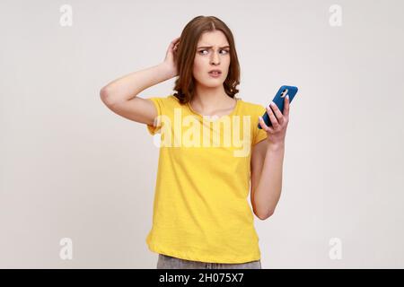 Pensive pensive fille de l'adolescence pensive en jaune style décontracté T-shirt debout avec téléphone mobile, touchant la tête, ayant doute sentiment de suspicion.Prise de vue en studio isolée sur fond gris. Banque D'Images