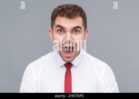 Portrait d'un jeune beau homme choqué ou surpris en chemise blanche et cravate debout avec le visage choqué, la bouche ouverte et regardant la caméra. Intérieur isolé sur fond gris. Banque D'Images