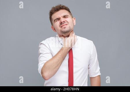 Douleur à la gorge.Portrait du jeune homme en chemise blanche et cravate debout et touchant son cou douloureux. Intérieur isolé sur fond gris. Banque D'Images