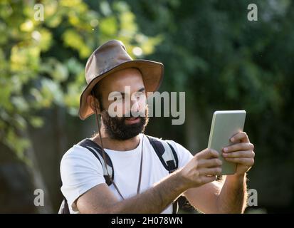 Jeune homme attrayant avec la barbe randonnée dans la forêt avec sac à dos sur les épaules et regardant la tablette Banque D'Images