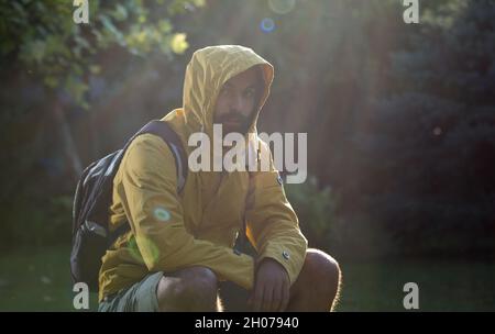 Jeune homme attrayant avec une barbe assise sur le rocher dans la forêt avec un imperméable et une capuche sur la tête Banque D'Images