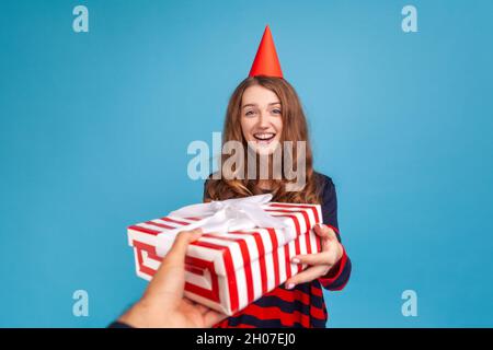 Femme positive portant un chandail à rayures style décontracté et un cône de fête rouge obtenant boîte de cadeau de personne inconnue, exprimant le bonheur, l'humeur festive.Studio d'intérieur isolé sur fond bleu. Banque D'Images