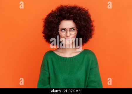 Portrait d'une femme confuse avec une coiffure afro portant un chandail vert de style décontracté et des lunettes optiques, regardant loin, frognant visage, pensant.Studio d'intérieur isolé sur fond orange. Banque D'Images