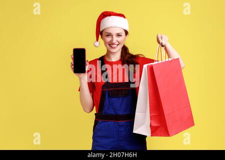 La femme de livraison montre un smartphone avec un écran vierge, tient des sacs d'achats avec des marchandises en ligne brasées, portant une combinaison bleue et chapeau de père noël.Studio d'intérieur isolé sur fond jaune. Banque D'Images