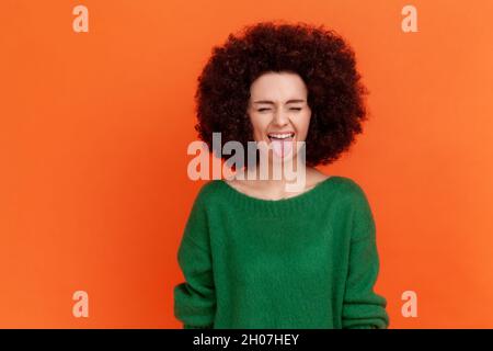 Positive joueur femme avec la coiffure afro portant le chandail vert debout avec les yeux fermés et la langue dehors, démontrant le comportement puéril.Studio d'intérieur isolé sur fond orange. Banque D'Images