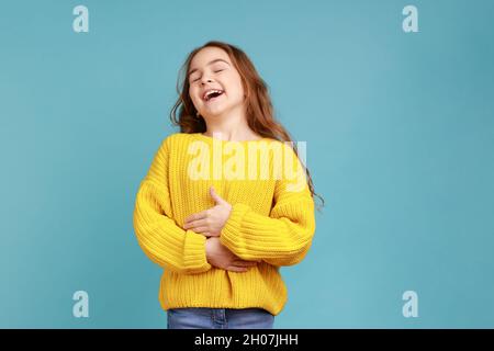 Portrait de bonne bonne bonne fille positive rire dehors fort, tenant son ventre, entendre drôle de blague, portant jaune style décontracté pull.Studio d'intérieur isolé sur fond bleu. Banque D'Images