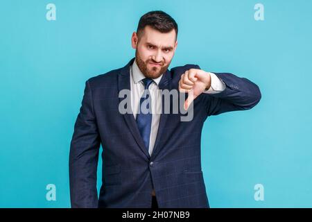 Portrait d'un homme barbu insatisfait portant une combinaison de style officiel montrant le pouce vers le bas et regardant l'appareil photo, n'aime pas, les commentaires de test d'application.Studio d'intérieur isolé sur fond bleu. Banque D'Images