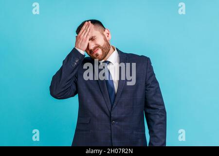 Portrait d'un homme barbu portant un costume de style officiel debout avec geste de facipalm, se blâmant lui-même, se sentant regret de tristesse à cause de la mauvaise mémoire.Studio d'intérieur isolé sur fond bleu. Banque D'Images