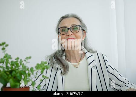 Femme d'affaires grise sénior vêtue d'une veste rayée à lunettes travaille dans son bureau assis au bureau. Bonne retraite, emploi et Banque D'Images
