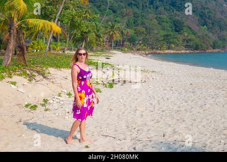 Une femme mûre avec de longs cheveux blonds marche le long d'une plage de sable parmi les palmiers sur une île tropicale en Thaïlande. Banque D'Images