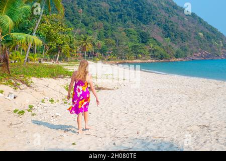 Une femme aux longs cheveux blonds marche le long de la plage de sable parmi les palmiers sur une île tropicale en Thaïlande. Banque D'Images