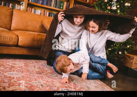 Thème des fêtes de Noël et du nouvel an en famille avec un petit enfant.Une famille de trois personnes, enveloppée dans une couverture, s'assoit sur le sol dans le salon près Banque D'Images