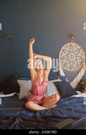 Femme paresseuse en pyjama élégant s'étire sur un lit confortable avec ordinateur portable Banque D'Images