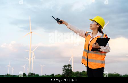 jeune femme ingénieur utilisant talkie walkie et le presse-papiers de lecture pour vérifier le système contre la ferme éolienne Banque D'Images