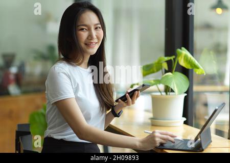 Jolie jeune femme asiatique tenant un téléphone mobile et un stylet, un ordinateur tablette sur la table dans le café Banque D'Images