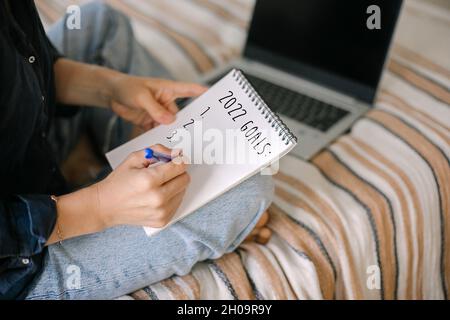 Jeune femme écrivant 2022 buts, à faire la liste, les plans tout en travaillant en ligne sur ordinateur portable de la maison.Concept de planification de la nouvelle année. Banque D'Images