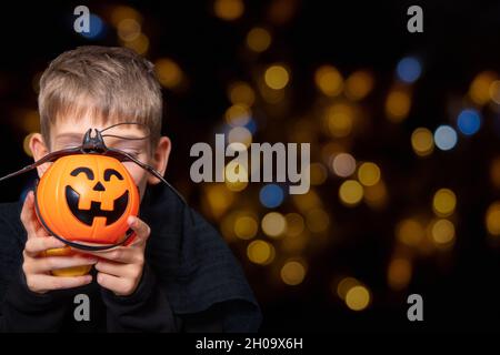 Un enfant tenant un panier orange en forme de citrouille avec un visage de repas, une lanterne de Jack et une chauve-souris sur un fond noir avec bokeh. Le garçon attend pour Banque D'Images