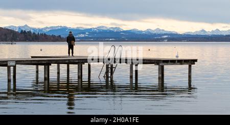 Herrsching, Allemagne - 22 janvier 2021: Homme debout sur un quai, regardant vers les alpes au loin. Au lac Ammersee. Banque D'Images