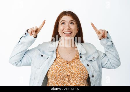 Bonne mignonne jeune femme avec des cheveux rouges, pointant et regardant avec le sourire heureux, en regardant les soldes d'halloween, le concept du vendredi noir, fond blanc Banque D'Images