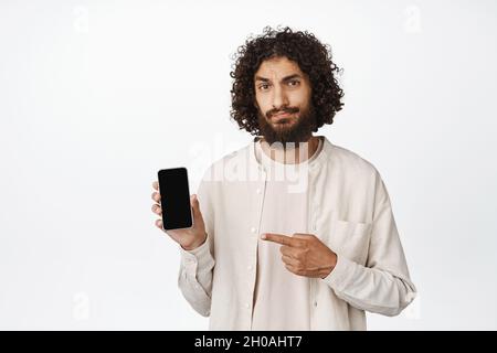 Jeune homme sceptique pointant vers l'écran de téléphone mobile, frognant et regardant douteux, n'aime pas l'application, fond blanc Banque D'Images