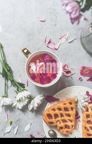 Pâtisserie en forme de coeur avec baies et fleurs roses thé dans une tasse de thé blanc sur table de cuisine en béton blanc avec fleurs.Un moment de thé confortable et romantique à la maison. Banque D'Images