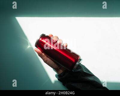 Bouteille Thermo-bidon rouge dans les mains mâles bleu turquoise mur fond ombre dure.Thé chaud café boissons boire en automne froid hiver temps.Dura Banque D'Images