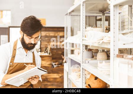 Jeune homme d'affaires séduisant un potter avec une barbe et une moustache travaille dans son atelier. Conserve les enregistrements et transcrites dans un ordinateur portable en inspectant Banque D'Images