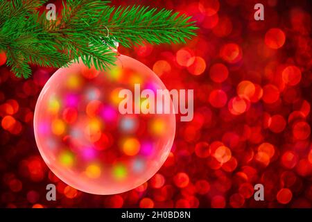 Boule de Noël transparente suspendue sur branche de pin vert sur fond de bokeh rouge. Banque D'Images