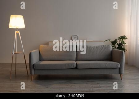 Canapé, horloge, plante en pot et lampe brillante le soir sur fond de mur gris dans le bureau ou le salon Banque D'Images