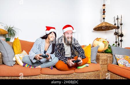 joyeux noël !Jeune couple adulte fêtant Noël en portant le chapeau du père noël assis sur le canapé et jouant ensemble des jeux vidéo. Famille Banque D'Images