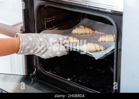 Mini croissants français traditionnels crus non cuits sur plaque de cuisson pour cuire au four.Petit déjeuner concept de boulangerie française.Main femelle dans le gant Banque D'Images