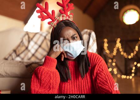 Jeune femme noire malheureuse portant des cornes de renne et un masque facial, célébrant Noël ou nouvel an seul à la maison Banque D'Images