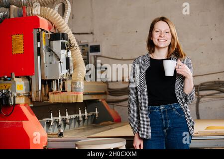 Une femme de travail tricherie vêtue de vêtements semi-officiels, debout à côté d'un gros morceau rouge de machinerie lourde.Elle tient une tasse de thé. Banque D'Images