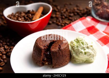 Une portion de fondant au chocolat avec une cuillère de glace au pistache sur une assiette.En arrière-plan se trouve une tasse en forme de cœur remplie de grains de café et d'un Banque D'Images