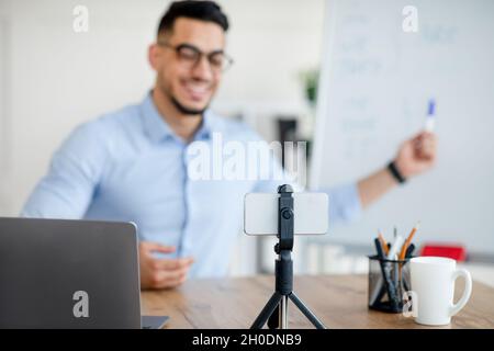 Joyeux professeur arabe de sexe masculin donnant une leçon d'anglais en ligne, en utilisant un smartphone sur trépied et ordinateur portable, mise au point sélective Banque D'Images