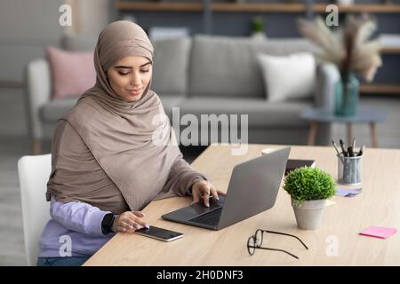 Femme musulmane assise à la table en utilisant la cellule et le pc Banque D'Images